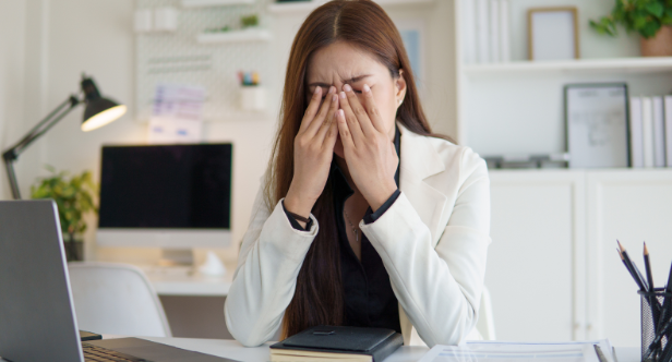 A young woman in an office covers her eyes from stress, illustrating the anxiety and sensory overload autistic students often face